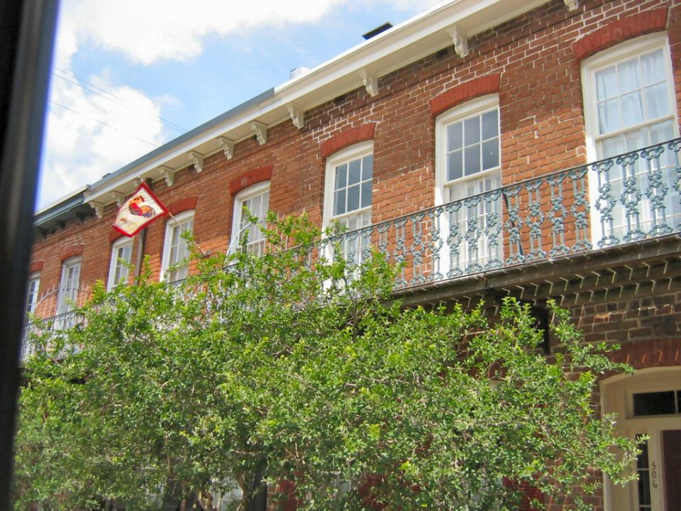 brick building with white windows and ornate balcony, green tree in foreground, flag with rooster emblem visible