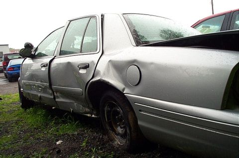 2001 Ford Crown Victoria - silver four-door sedan showing heavy collision damage along the driver side, parked on dirt by other cars.