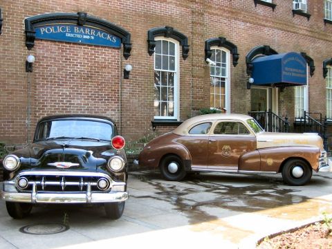1985 Ford LTD Crown Victoria - Two vintage police cars are parked in front of a brick police barracks building.