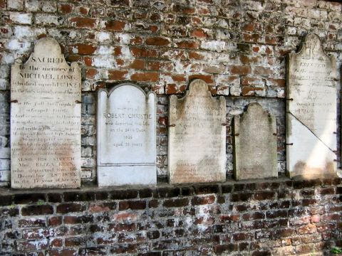 1985 Ford LTD Crown Victoria - A row of old weathered headstones set into an aged brick wall, with partially legible inscriptions memorializing individuals including Mich…
