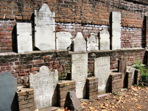 1985 Ford LTD Crown Victoria - A row of weathered antique gravestones from the early 1800s set against and along an old red brick wall, with fallen leaves on the ground.