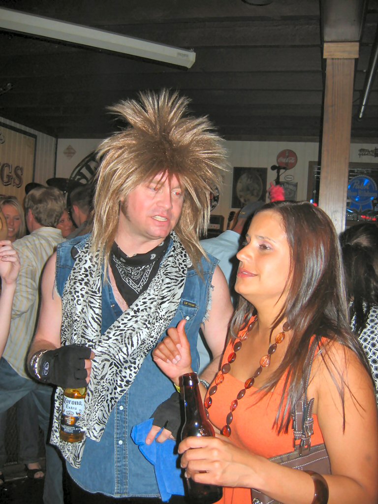 Man in a blonde wig and denim vest talks to a woman in an orange top at a bar