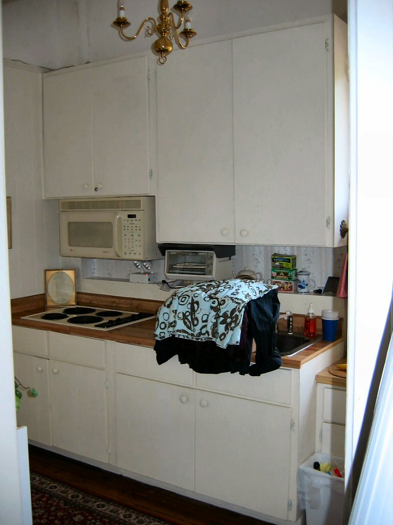 Kitchen counter with a stovetop, microwave, sink, and cabinets. Clothes are piled on the counter