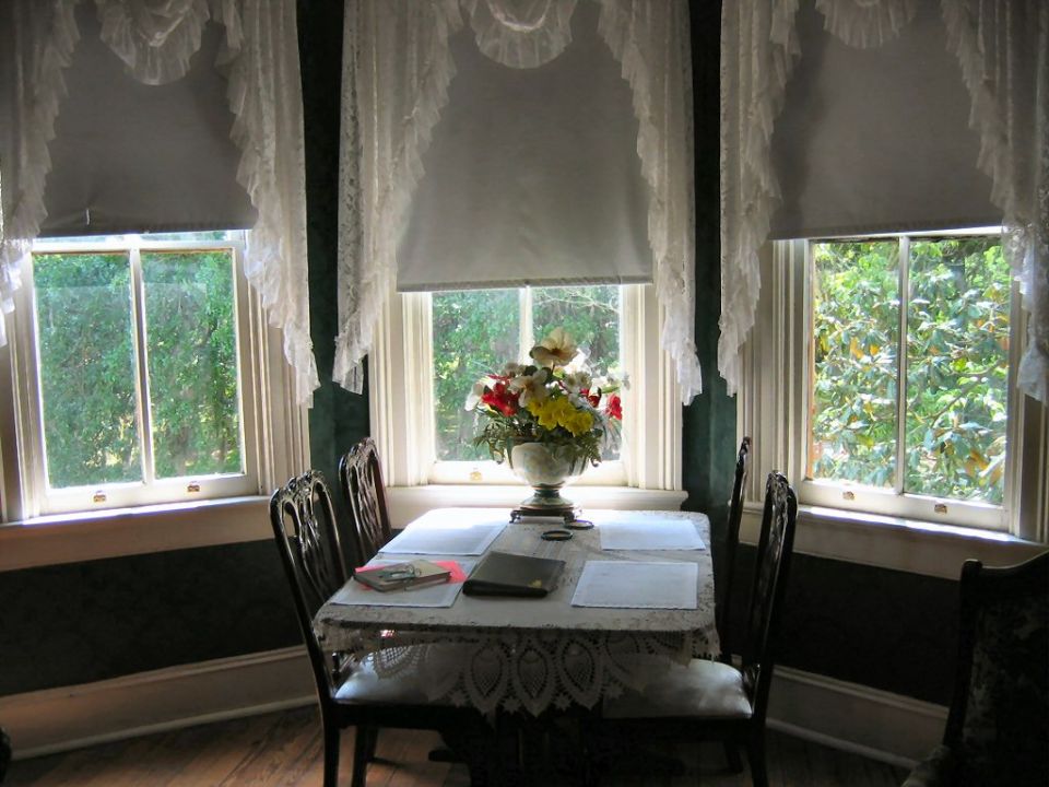 Dining room table set with papers and a floral centerpiece, viewed through lace-curtained windows