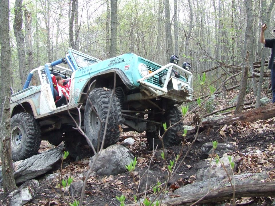 1969 Ford Bronco off-roading on a rocky trail, climbing over large boulders. Blue and white paint, roll cage, and large tires visible