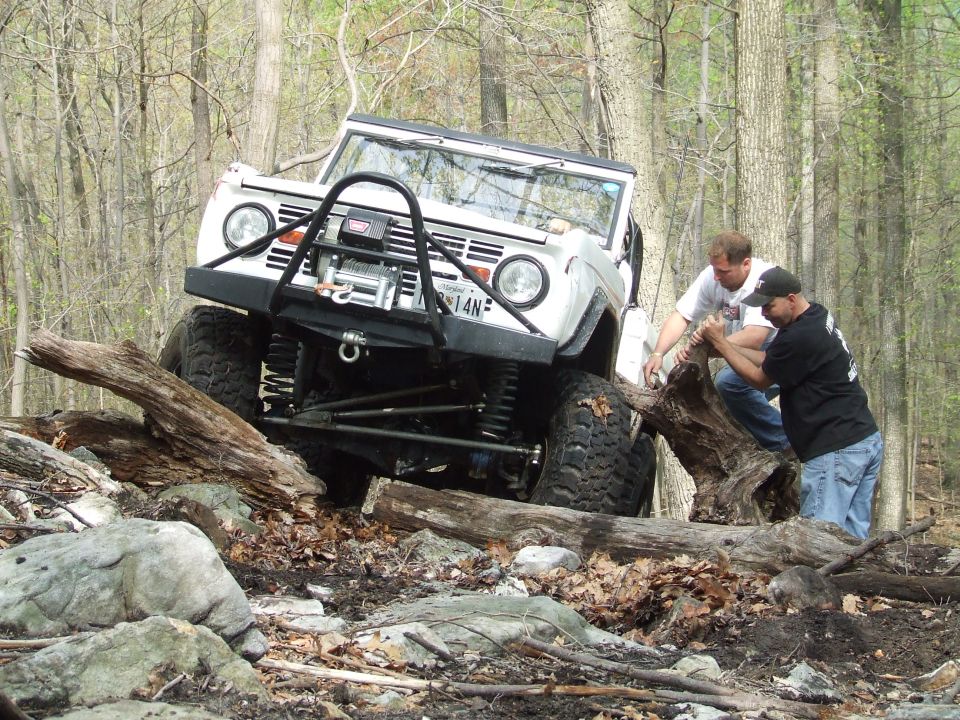 White off-road SUV with winch on a rocky, wooded trail. Two men are pushing a log near the front tire