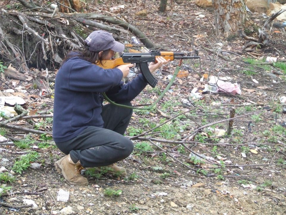 Person in a blue hoodie and baseball cap crouches in woods, aiming an AK-47 rifle