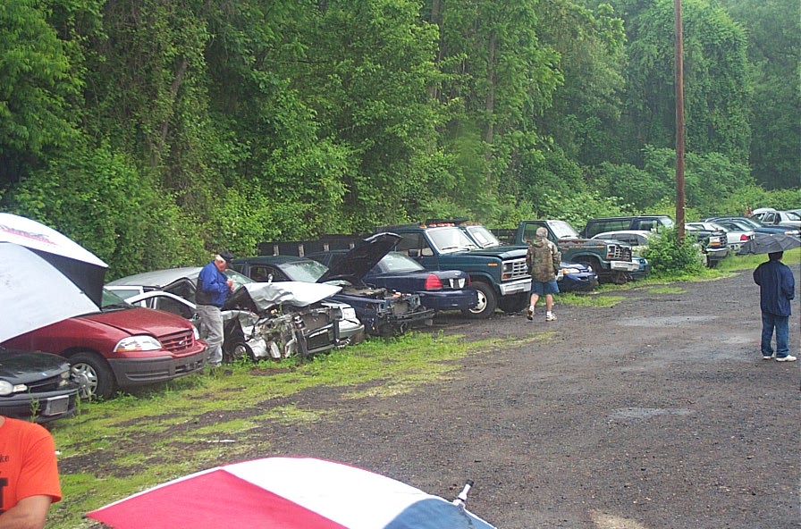 Cars parked in a lot with trees in the background. Some cars have hoods up