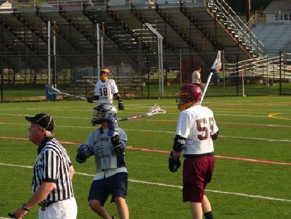 Lacrosse players and referee on a field with stadium seating
