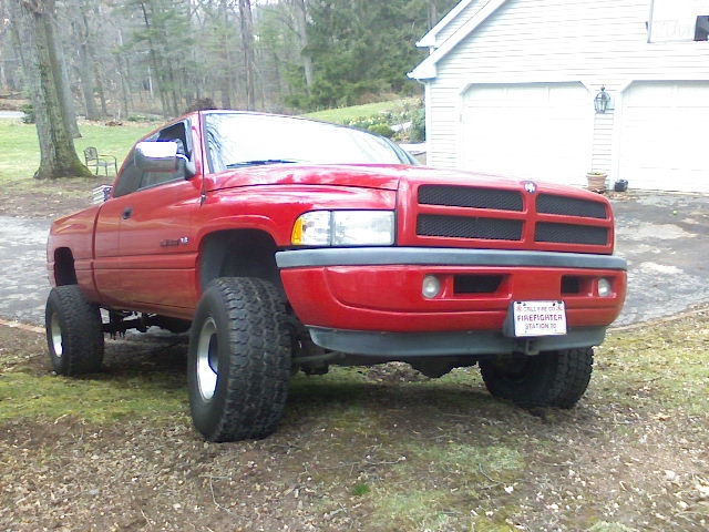 Red 1997 Dodge 1500 pickup truck with lifted suspension and large tires, parked on grass in front of a garage