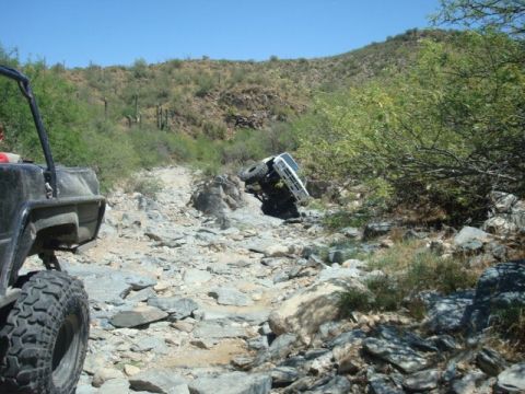 1986 Toyota Hilux - A white 4x4 climbs off-camber up a boulder-filled desert wash, front tire lifted and body tilted hard to one side, while a second rig with…