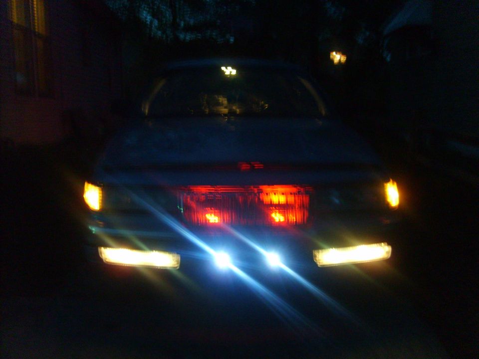 Car front at night, headlights and turn signals illuminated. Red taillights visible