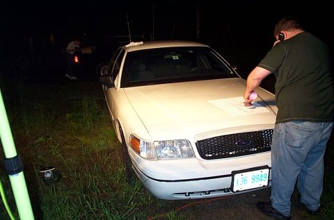 2001 Ford Crown Victoria - A man talks on his phone while reviewing a document placed on the hood of a white Ford Crown Victoria at night.