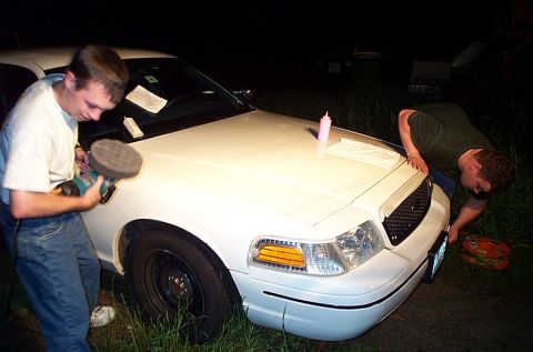 2001 Ford Crown Victoria - Two people buff and detail a white Ford Crown Victoria at night outdoors, one holding a rotary polisher with a foam pad at the front fender…