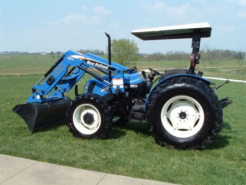 1984 Honda ATC 250 R - A blue New Holland tractor with a front loader is parked on a grassy field under a bright blue sky.