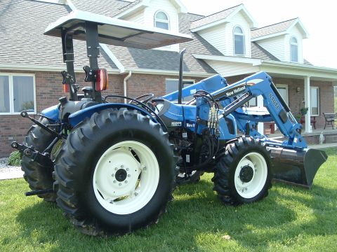 1984 Honda ATC 250 R - A blue New Holland tractor with a front loader is parked on a grassy lawn in front of a brick house.