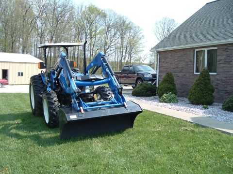 1984 Honda ATC 250 R - A blue tractor equipped with a front loader is parked on a grassy lawn, with a house and a dark pickup truck visible in the background.