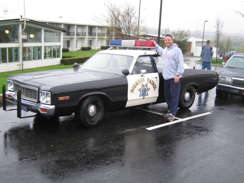 Black and white CHP Highway Patrol classic sedan in wet parking lot, man standing beside it