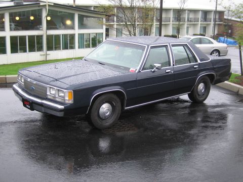 1991 Ford Crown Victoria - A dark blue 1991 Ford Crown Victoria sedan is parked on a wet surface, with a building in the background.