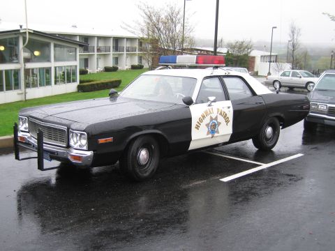 1991 Ford Crown Victoria - This black and white highway patrol car, likely a 1977 model, features a push bumper and a light bar on the roof.