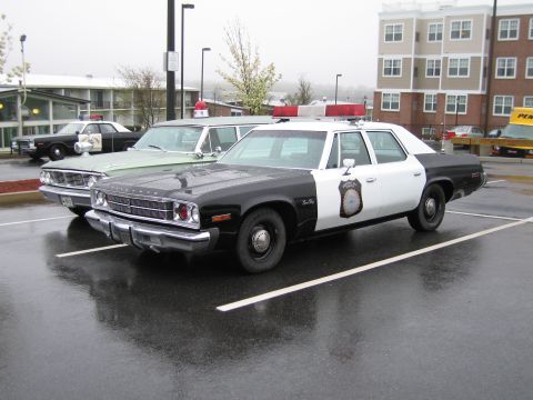 1991 Ford LTD Crown Victoria - black-and-white Plymouth police sedan with rooftop lightbar parked on wet pavement, a green sedan behind it.