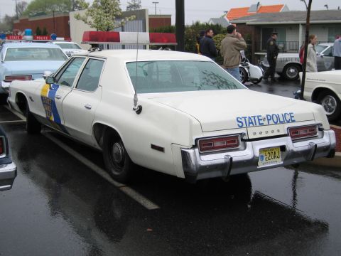 1991 Ford LTD Crown Victoria - white Dodge State Police sedan with a blue and gold side stripe viewed from the rear among other classic cruisers.