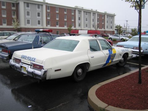1991 Ford LTD Crown Victoria - white Dodge State Police sedan with blue and gold stripe parked on wet pavement near a brick building.