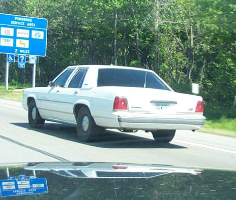2001 Ford Crown Victoria - A white sedan drives down the highway, passing a service area sign.