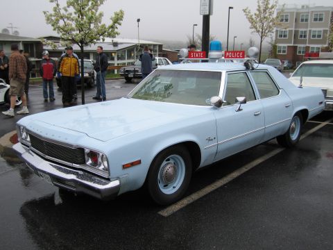 1991 Ford LTD Crown Victoria - light blue Plymouth State Police sedan with a roof lightbar parked on wet pavement as spectators stand nearby.