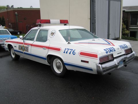 1991 Ford LTD Crown Victoria - white State Police sedan with red-white-blue stripes and the number 1776 viewed from the rear by a building.
