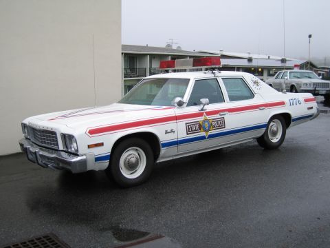 1991 Ford LTD Crown Victoria - white State Police sedan with red-white-blue stripes, a star shield, and the number 1776 parked by a building.