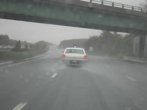 1991 Ford LTD Crown Victoria - Seen through a rain-spotted windshield, a cream police car with a roof beacon drives a wet highway beneath an overpass.