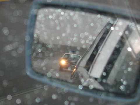 1991 Ford LTD Crown Victoria - car side mirror, dotted with rain, reflecting a police car with one headlight on following behind on a wet road.