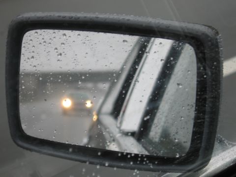 1991 Ford LTD Crown Victoria - rain-covered side mirror reflecting an oncoming car with headlights on against a gray highway backdrop.