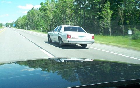 2001 Ford Crown Victoria - A white 2001 Ford Crown Victoria sedan is seen from behind on a highway surrounded by trees.