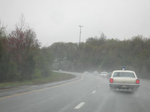 1991 Ford LTD Crown Victoria - cream police car with a blue roof beacon drives a misty wet highway with scattered traffic and roadside trees.