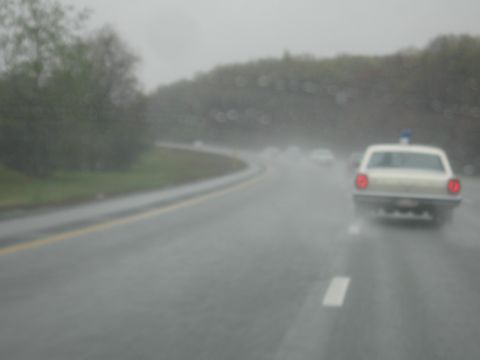 1991 Ford LTD Crown Victoria - blurry cream police car with a blue roof beacon and glowing taillights drives a rain-soaked highway curve.