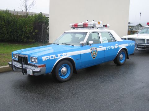 1991 Ford LTD Crown Victoria - blue-and-white NYPD Police Dodge sedan with blue wheels and twin red roof beacons parked beside a building.