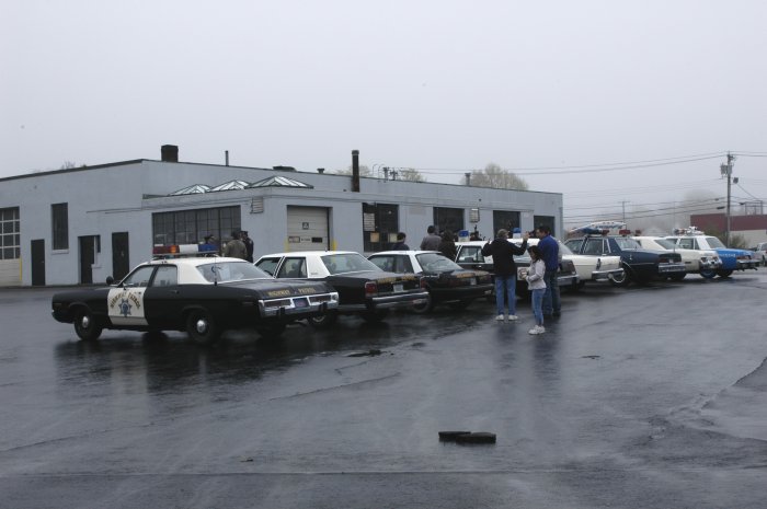 Police cars parked in a lot on a cloudy day. A black and white car is in the foreground with "Sheriff" text visible