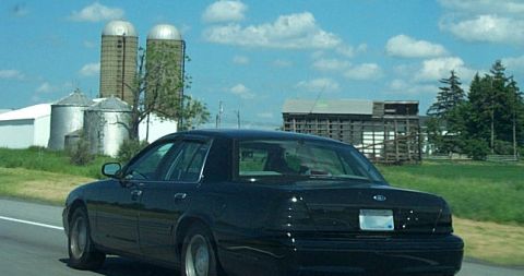 2001 Ford Crown Victoria - Rear view of a black 2001 Ford Crown Victoria on a highway, with farm buildings under a cloudy sky.