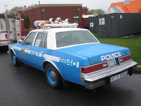 1991 Ford LTD Crown Victoria - blue-and-white New York City police Dodge sedan parked beside a fire engine, marked 'POLICE 2840' and 'OUT OF SERVICE' on a wet lot.