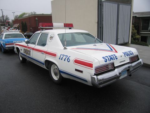 1991 Ford LTD Crown Victoria - white 1970s full-size State Police sedan numbered 1776 with red, white and blue stripes parked on a wet lot, another police car behind it.