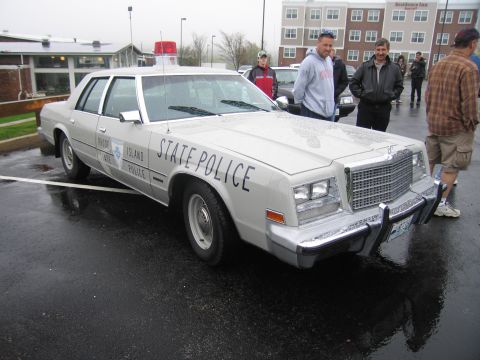 1991 Ford LTD Crown Victoria - silver Chrysler State Police sedan parked on a wet lot with several men standing nearby, building in the background.