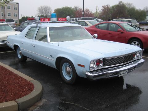 1991 Ford LTD Crown Victoria - light-blue 1970s Dodge four-door sedan fitted with a police roof lightbar parked among other cars on a wet lot.