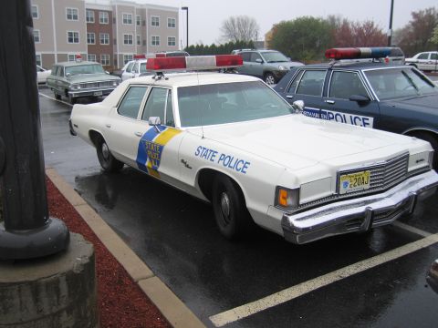 1991 Ford LTD Crown Victoria - white Dodge State Police sedan with a blue-and-yellow door emblem and roof lightbar parked beside other patrol cars on a rainy lot.