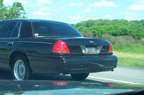 2001 Ford Crown Victoria - Rear view of a black 2001 Ford Crown Victoria on a highway with trees in the background.