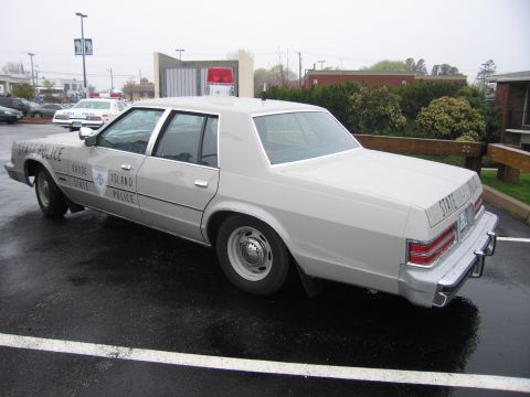 1991 Ford LTD Crown Victoria - tan Rhode Island State Police sedan with a door shield and roof lightbar parked on a wet lot beside other patrol cars.