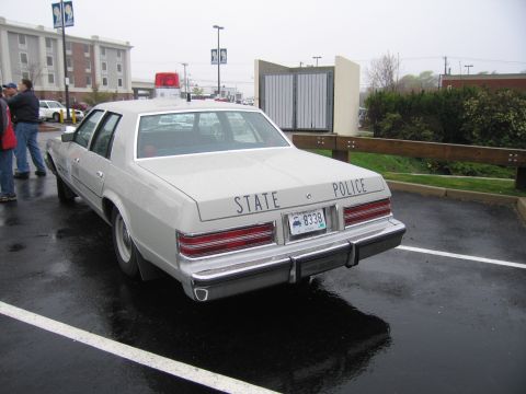 1991 Ford LTD Crown Victoria - cream full-size State Police sedan, rear view, with 'STATE POLICE' lettering across the trunk and a roof lightbar on a wet lot.
