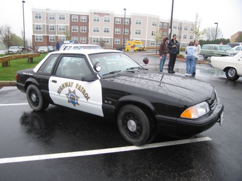 1991 Ford LTD Crown Victoria - black-and-white California Highway Patrol Ford Mustang 5.0 coupe parked on a wet lot, people standing nearby.