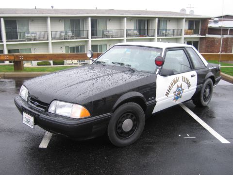 1991 Ford LTD Crown Victoria - black-and-white California Highway Patrol Ford Mustang 5.0 coupe with door star emblem parked before a motel on a wet lot.